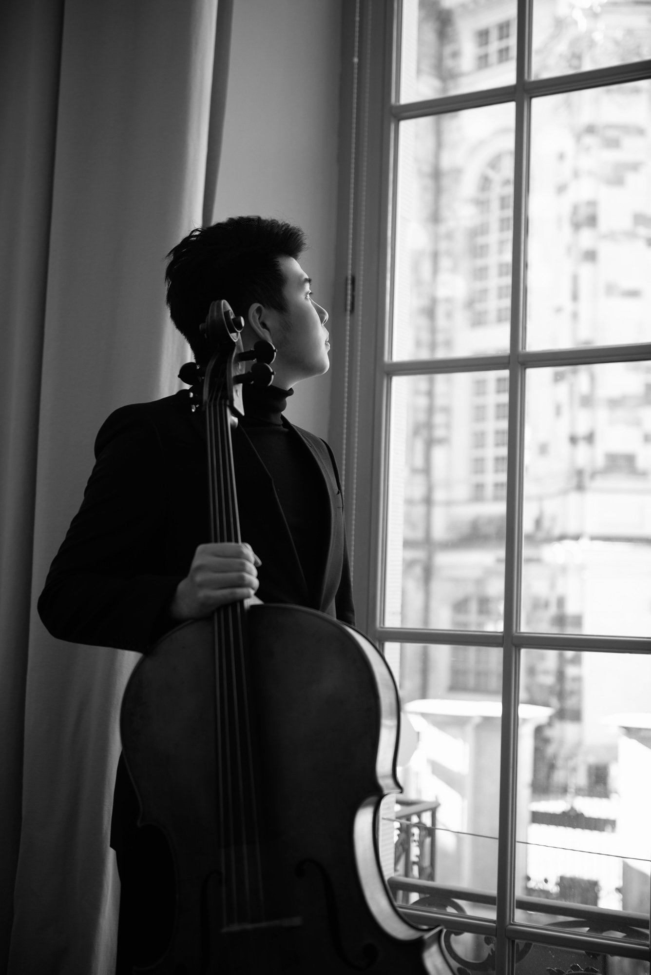 Black-and-white portrait of Yun Ho holding his cello and looking out of a window