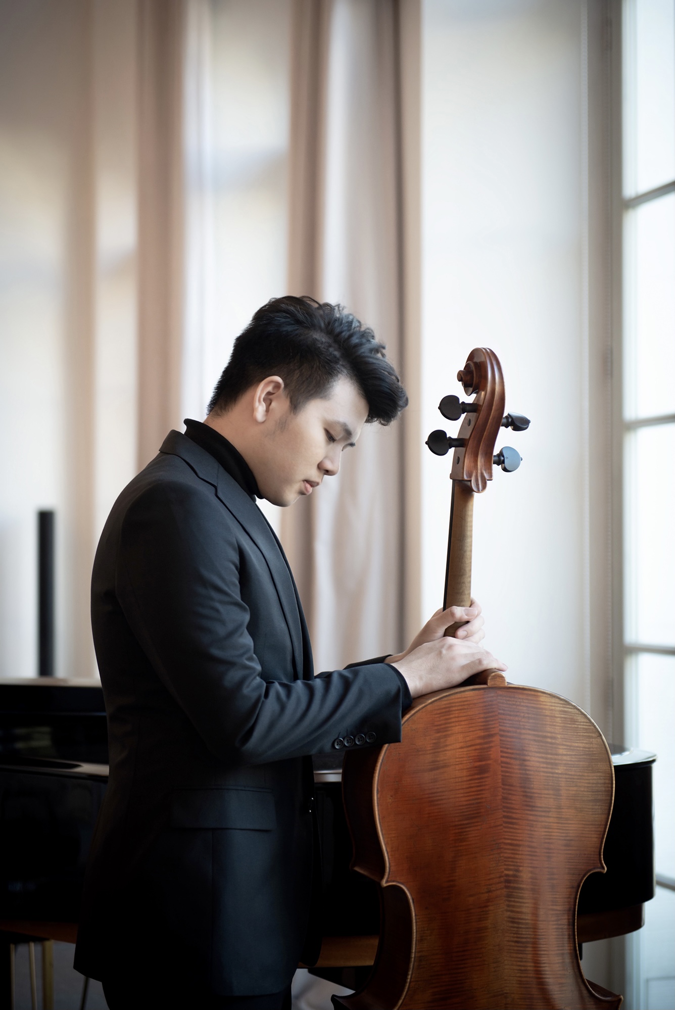 Yun Ho looking down at his cello beside a window and grand piano
