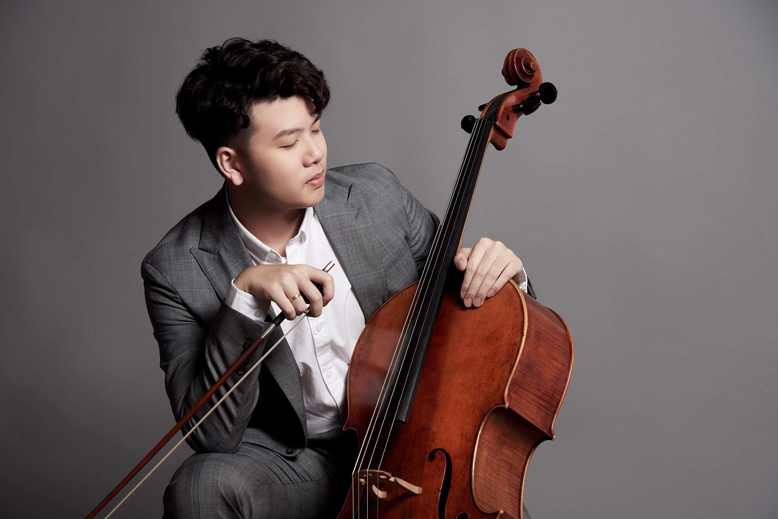Yun Ho seated with cello and bow in a grey studio portrait