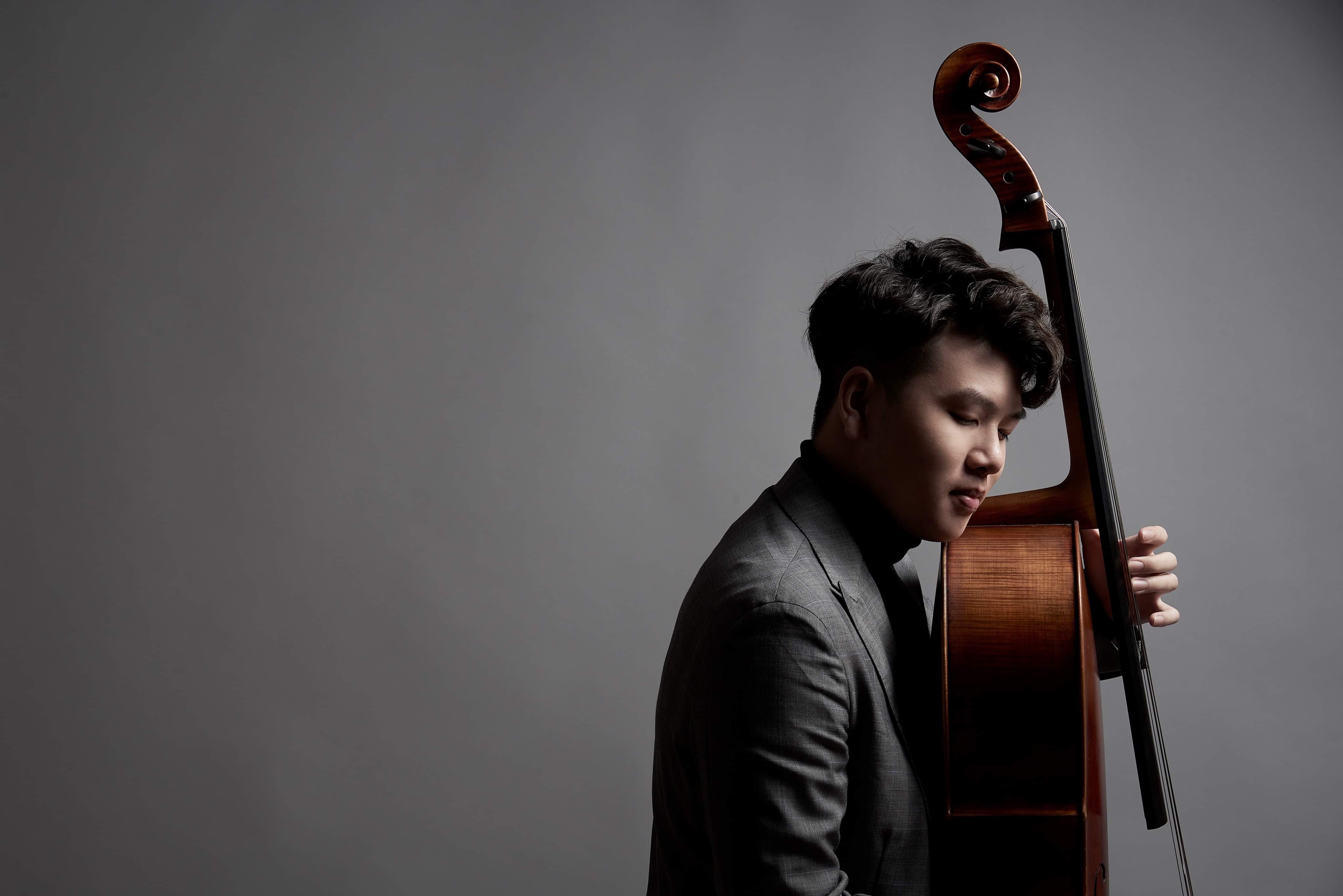 Yun Ho standing beside his cello in a grey studio portrait